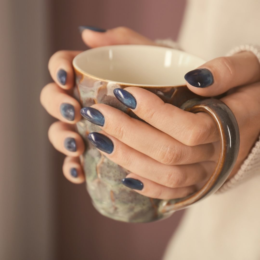 Hands with dark blue almond acrylic nails holding a marbled ceramic mug against a soft pink background