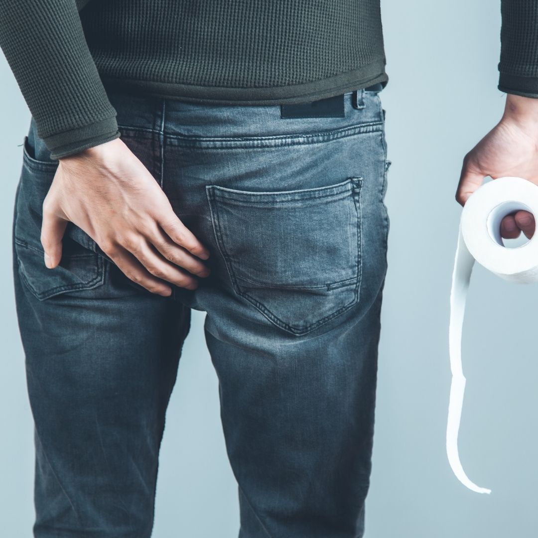 Person from behind touching their left buttock with left hand and holding a roll of toilet paper in their right hand