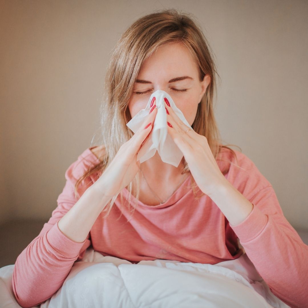 Woman blowing her nose into a tissue while sitting on a bed, eyes closed, wearing a pink long-sleeve top