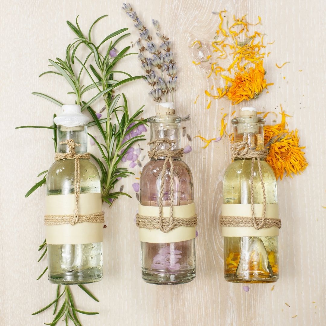 Three small glass bottles of essential oils with rosemary, lavender, and calendula petals on a light wood surface