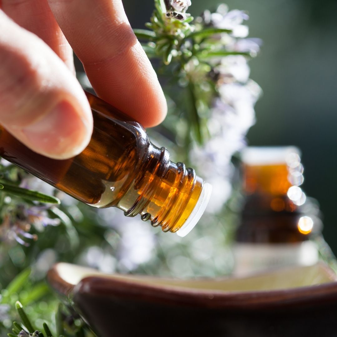 Hand tilting an amber essential oil bottle to pour a drop into a small ceramic dish, rosemary sprigs blurred in the background
