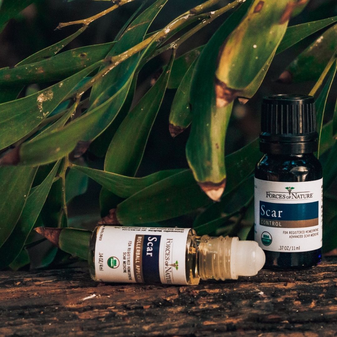 Close up of two Forces of Nature Scar bottles: rollerball lying on wood and small bottle standing in front of green leaves