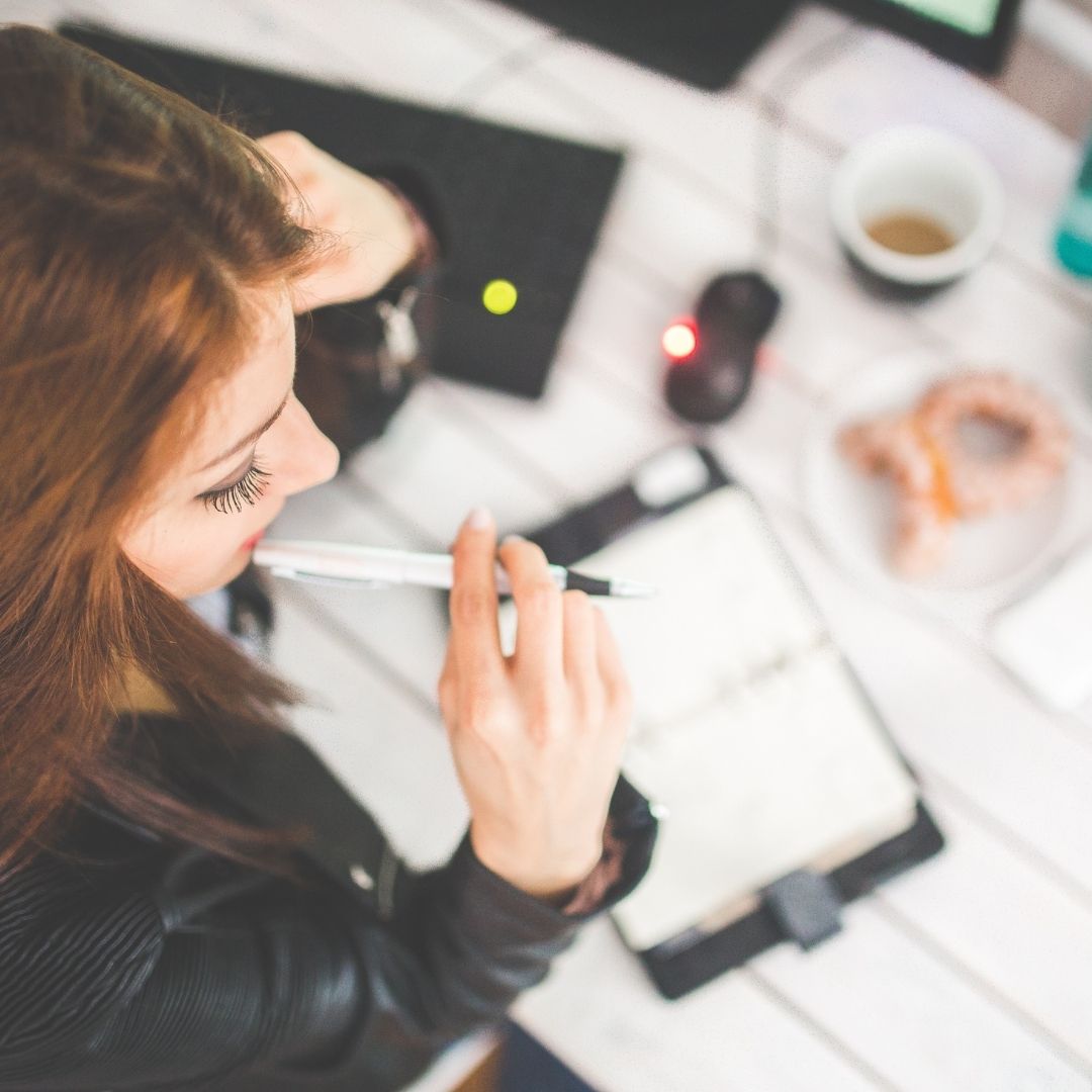 Woman holding a pen and thinking over an open planner at a cluttered desk with a coffee cup and pastry nearby