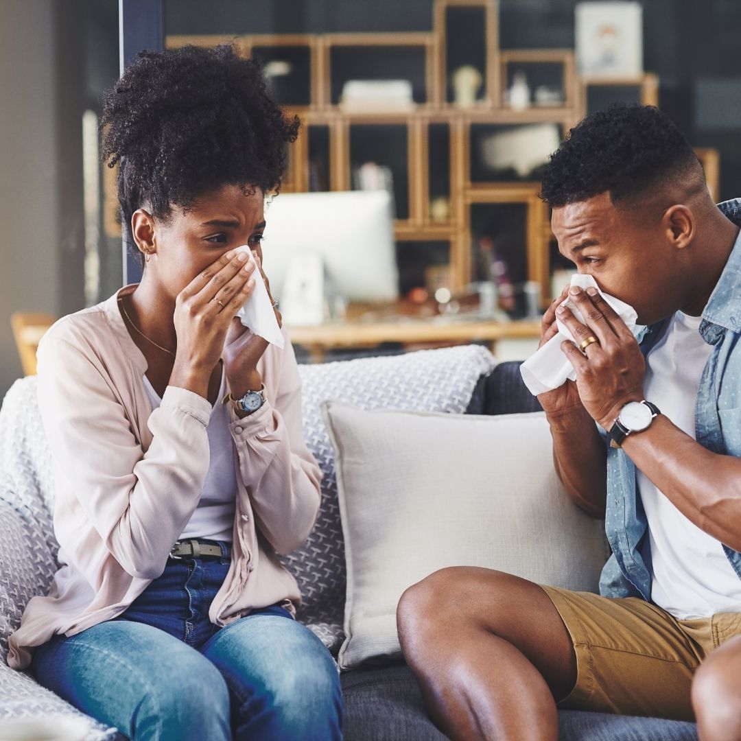 Man and woman sitting on a couch blowing their noses into tissues in a living room