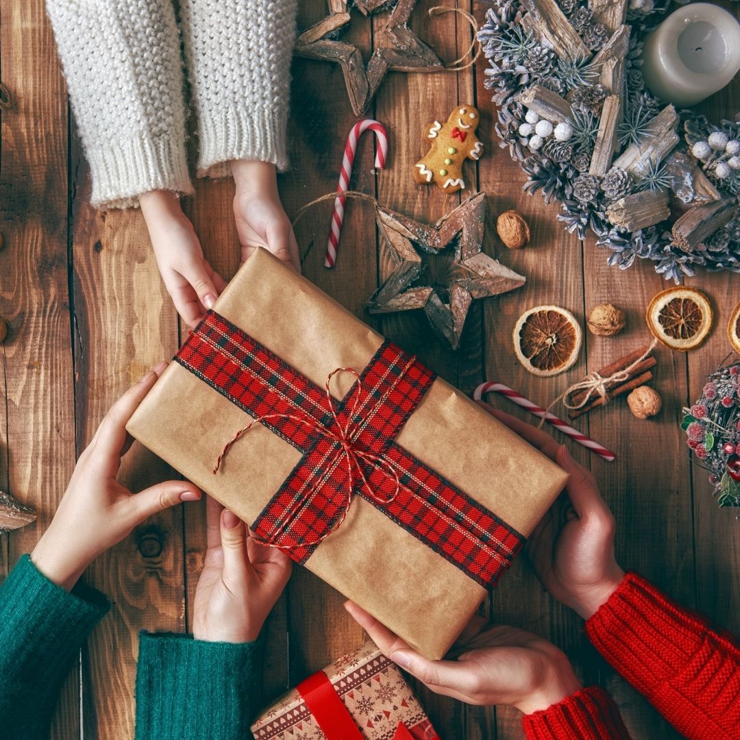 Four hands passing a wrapped gift over a wooden table with a holiday wreath, gingerbread cookie, candy canes, and dried orange slices