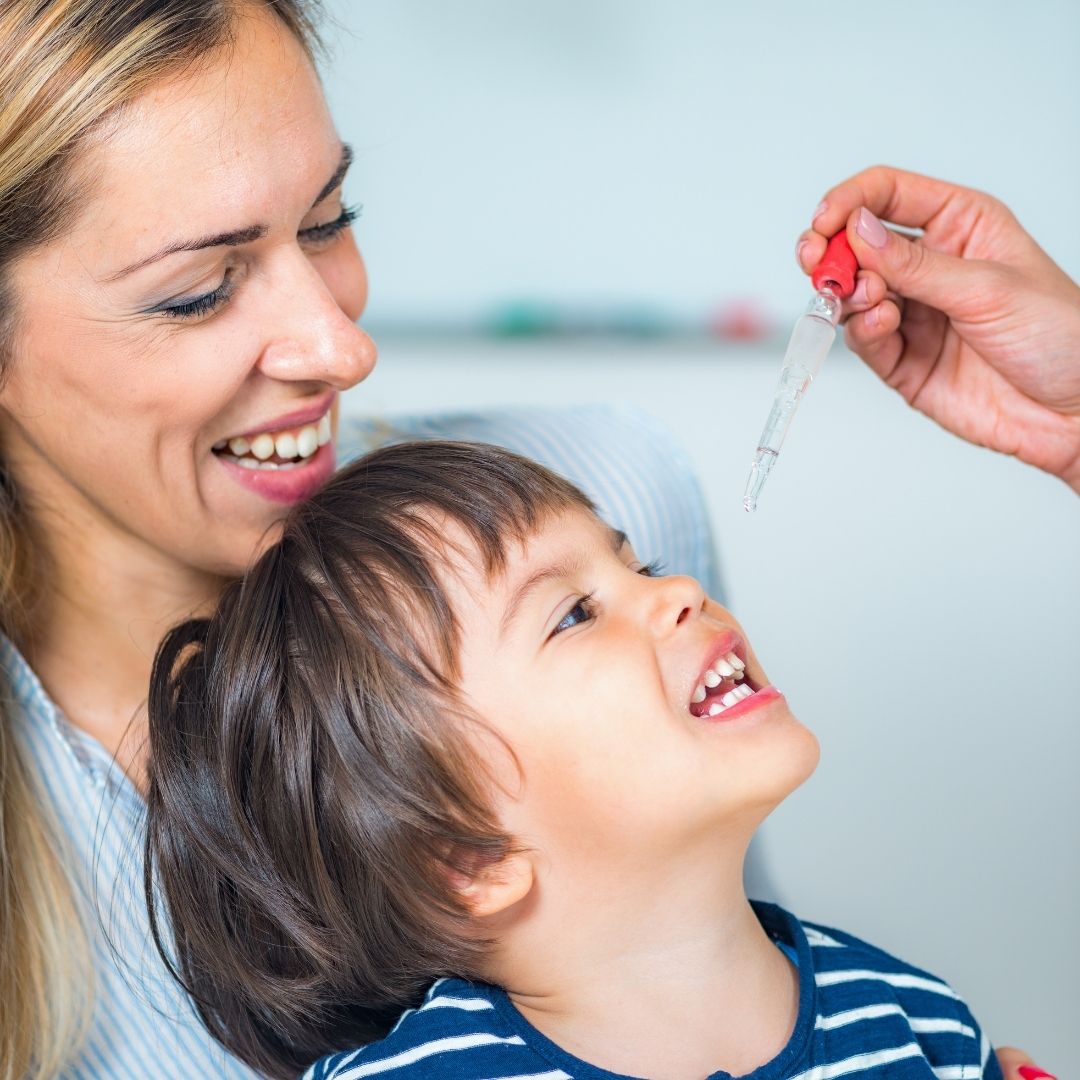 Woman holding a smiling toddler as a hand uses a dropper to give liquid to the child’s mouth in a clinic-like setting