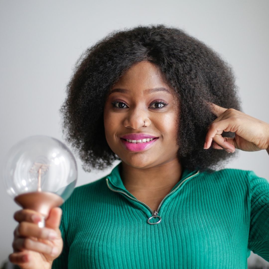 Smiling woman holding a clear light bulb and tapping her temple as if having an idea, neutral gray background