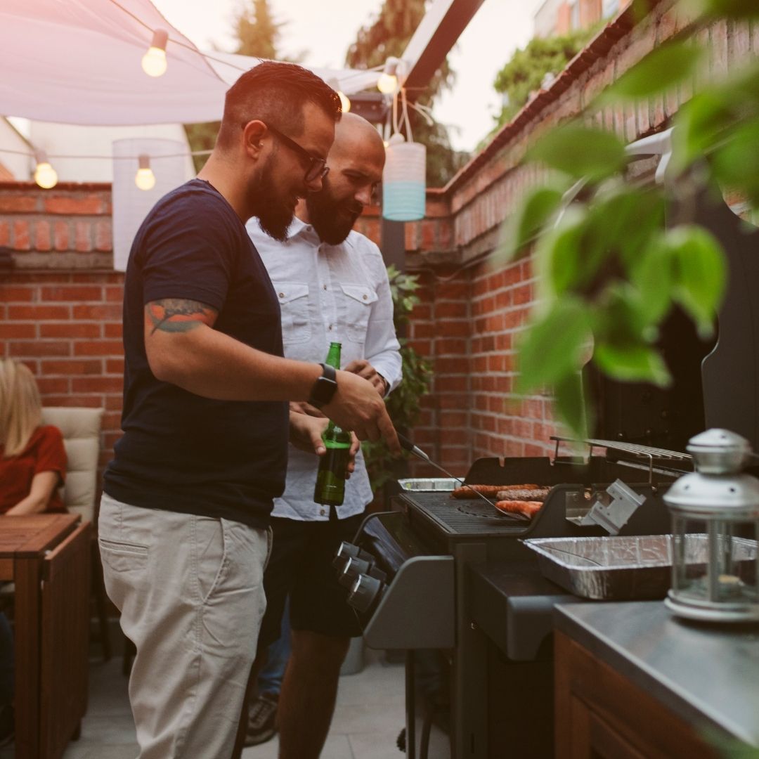 Two men grilling sausages on a backyard gas grill, one holding a beer bottle beneath string lights and a brick patio
