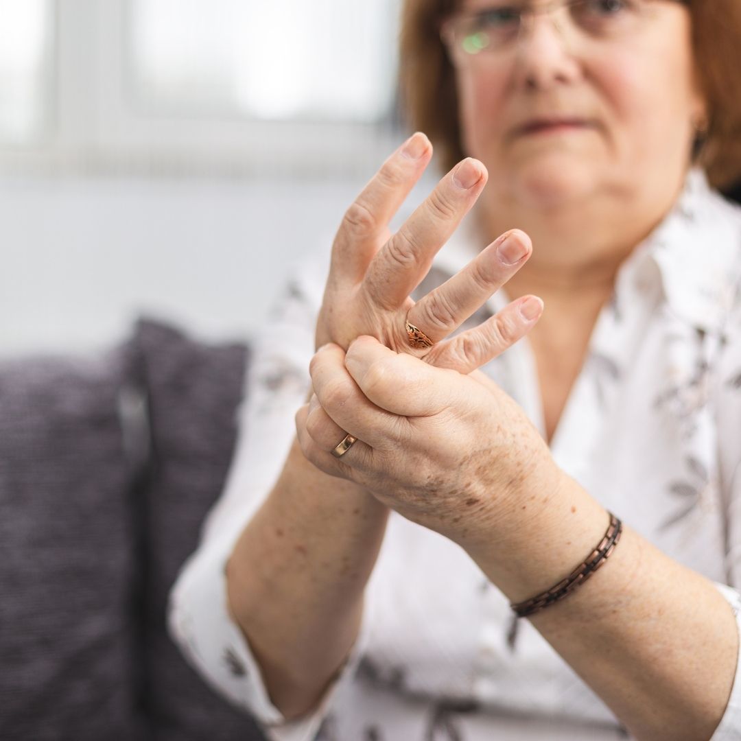 Older woman rubbing her hand and fingers as if easing joint pain while sitting on a couch