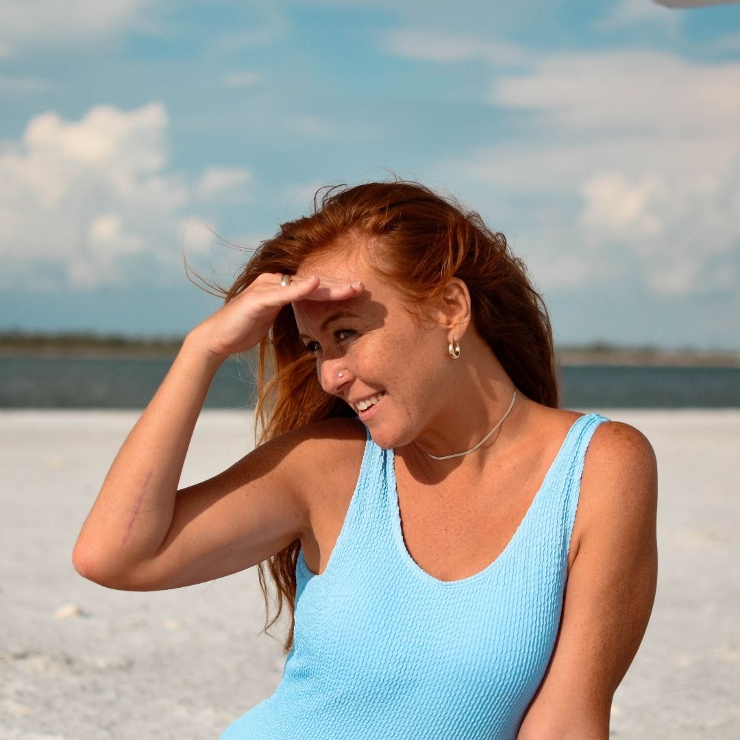 Red-haired woman shading her eyes and smiling on a sunny beach wearing a light blue tank top
