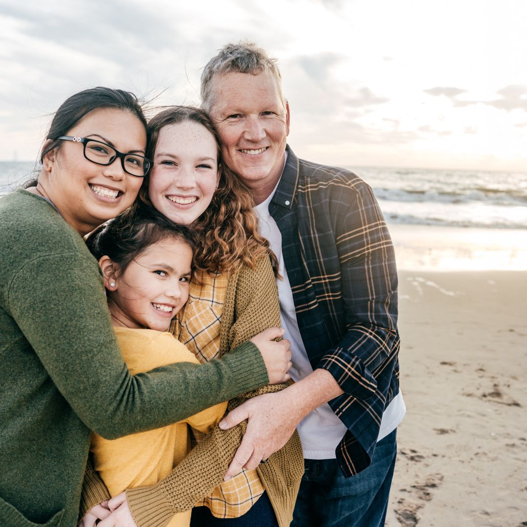 Two parents and two daughters hugging and smiling on a sandy beach at sunset