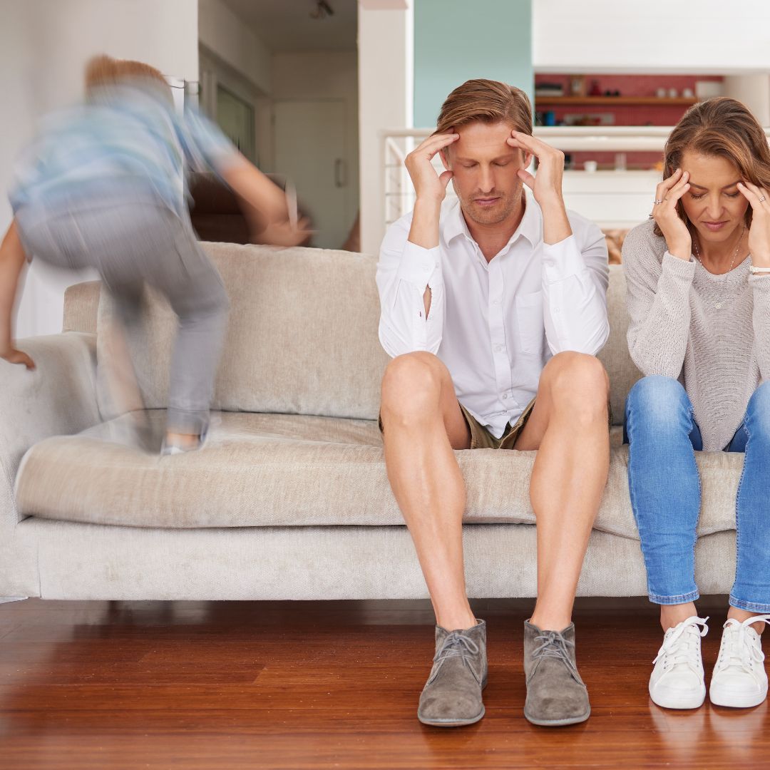 Parents sitting on a couch looking stressed while an energetic child jumps beside them in a living room