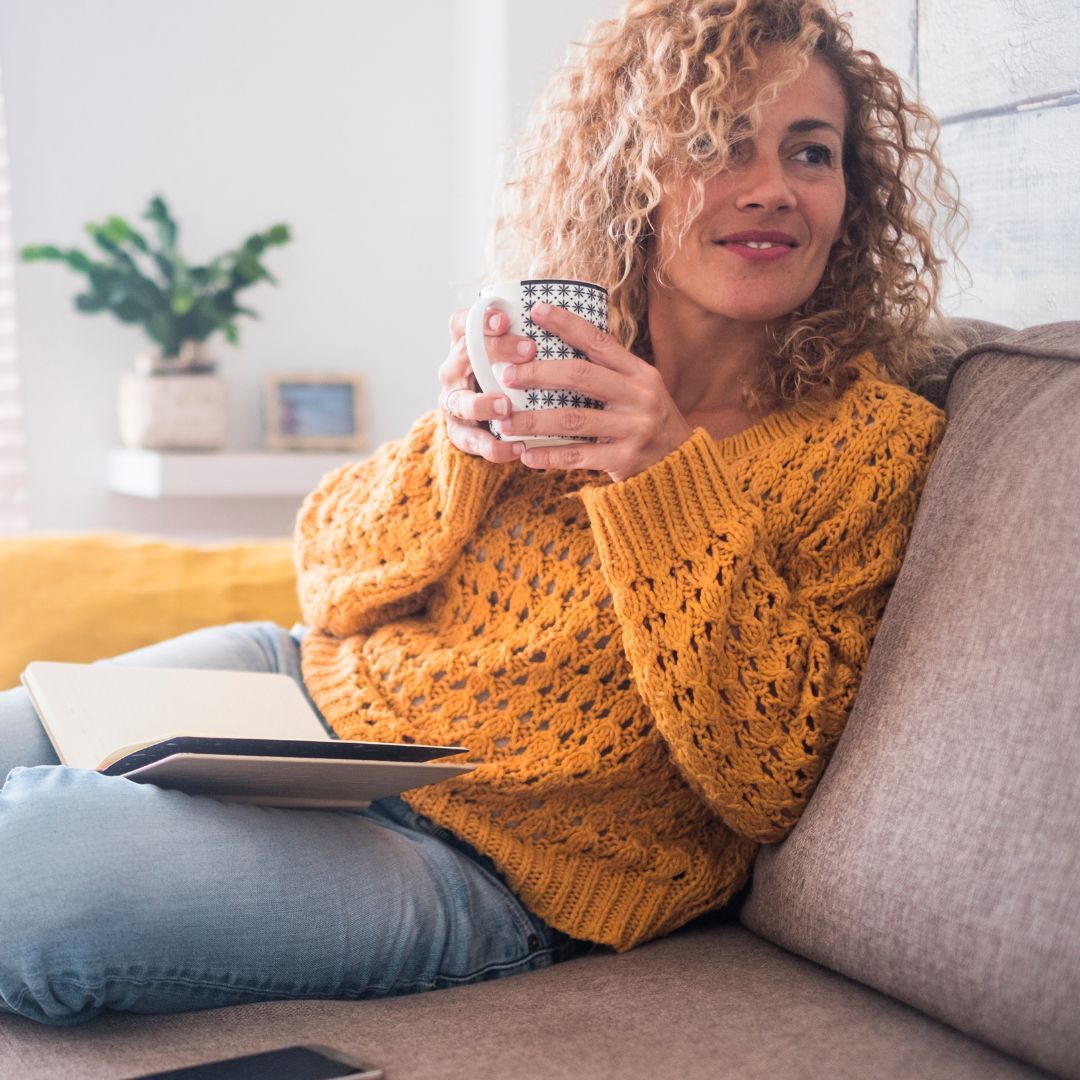 Woman sitting on a couch holding a patterned mug and an open notebook, wearing a mustard knit sweater in a bright living room