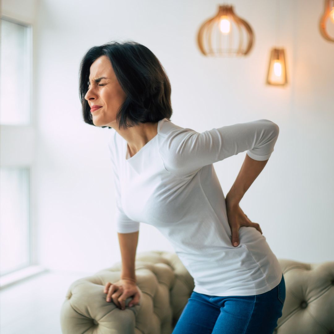 Woman clutching her lower back in pain while leaning on a sofa in a bright living room