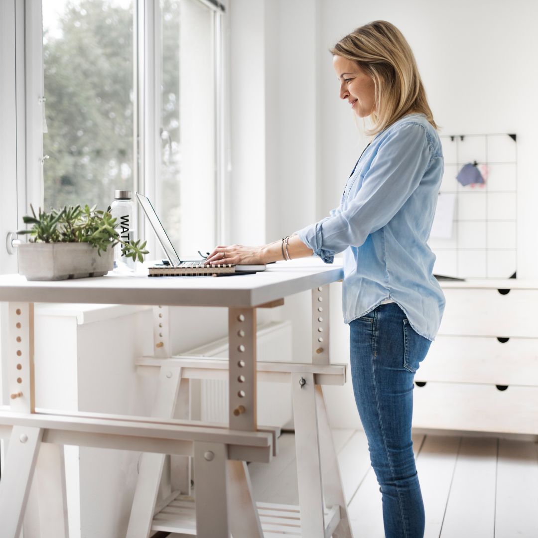 Woman standing at an adjustable standing desk typing on a laptop in a bright home office