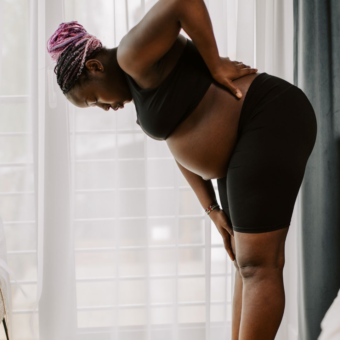 Pregnant woman bending forward and holding her lower back in pain by a sheer curtained window, wearing black top and shorts