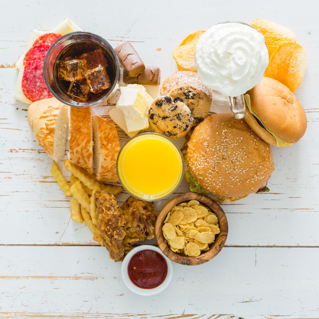 Variety of common acid reflux trigger foods on a white wooden table, including burger, fries, soda, orange juice, cookies