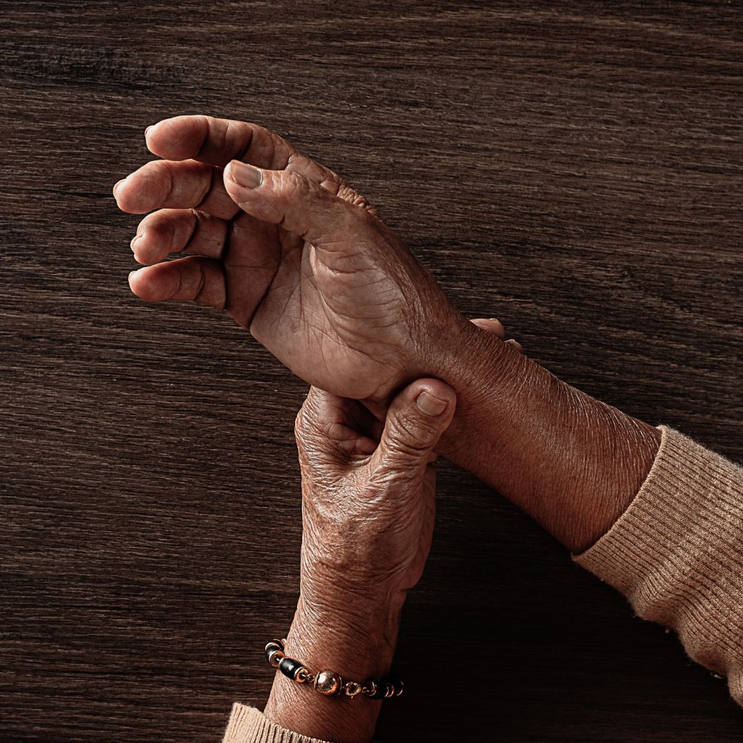 Older person’s hands, one hand holding the other wrist as if easing joint pain, on a dark wood table