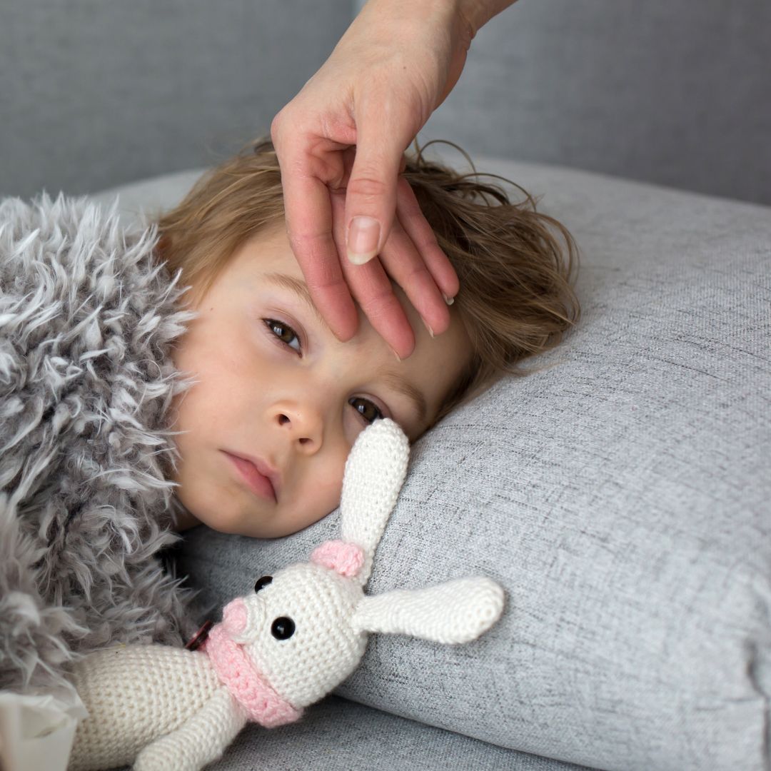 Child lying on a couch looking sick while an adult checks their forehead, knitted bunny tucked beside them