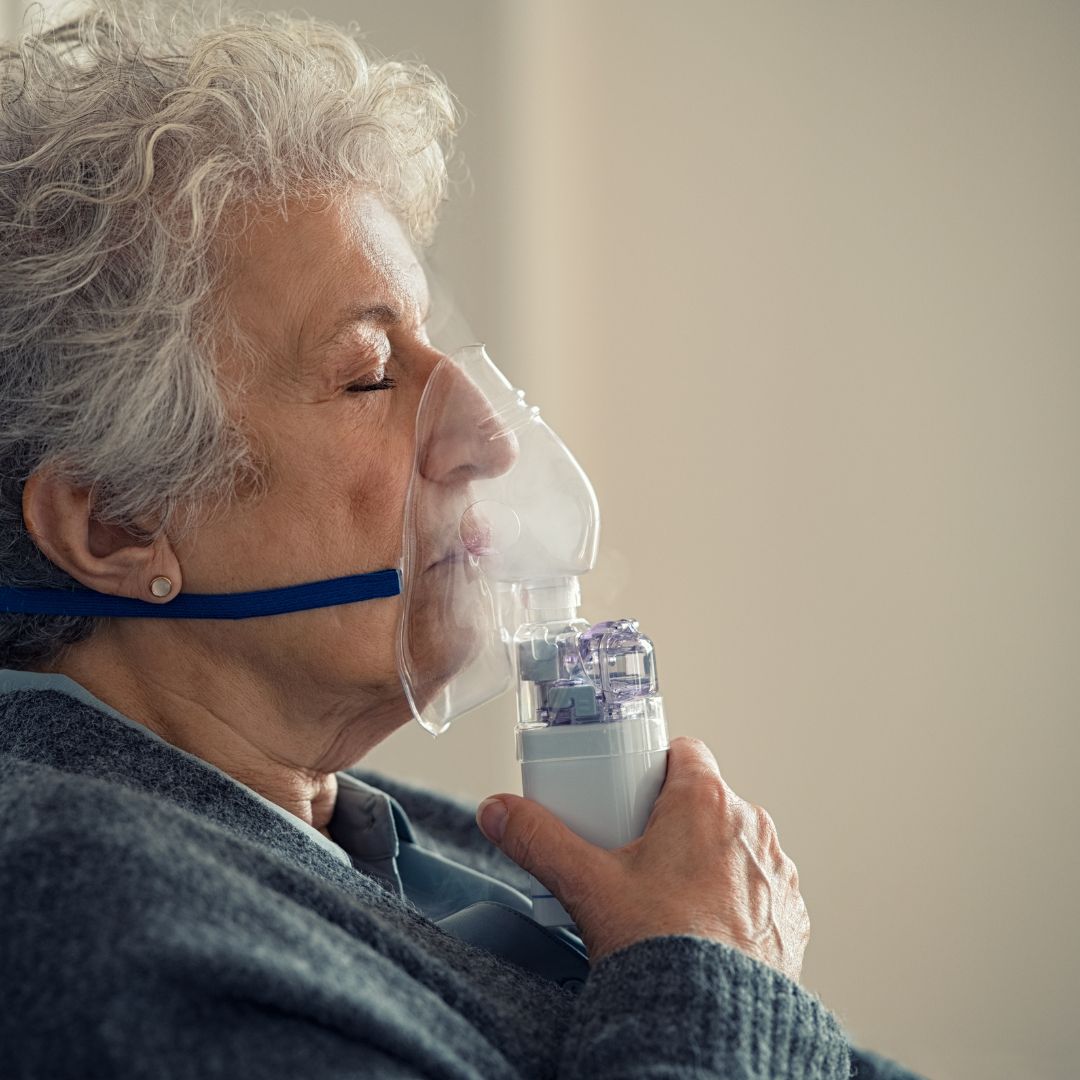 Elderly woman using a handheld nebulizer with a clear face mask at home, eyes closed