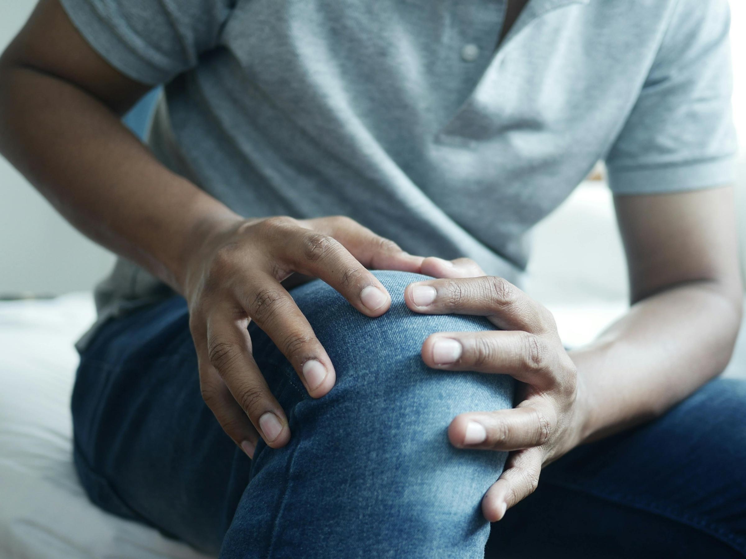 Person pressing their knee with both hands while seated on a bed, close-up showing jeans and a gray shirt