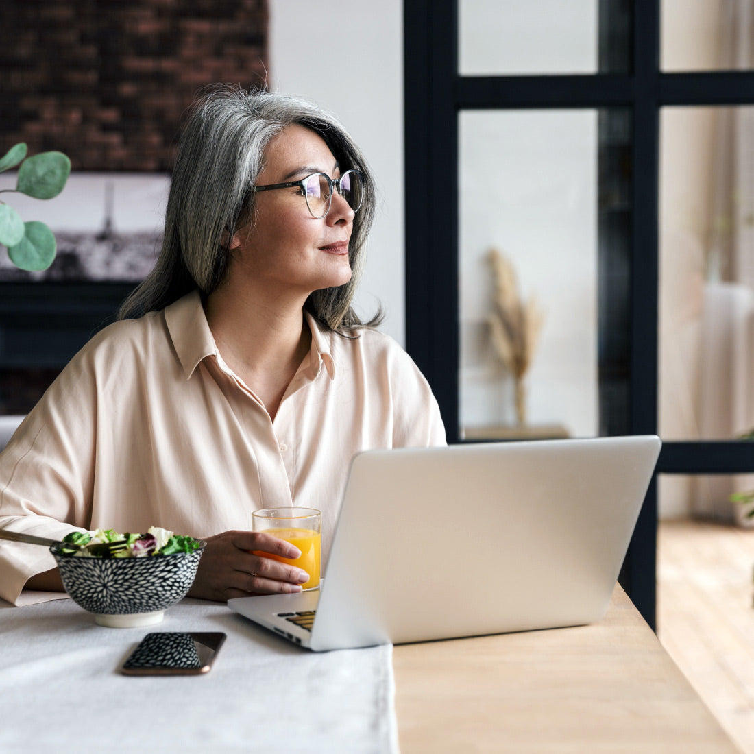 Older woman with gray hair and glasses enjoying breakfast while working on a laptop, symbolizing cognitive health, focus, and memory support.
