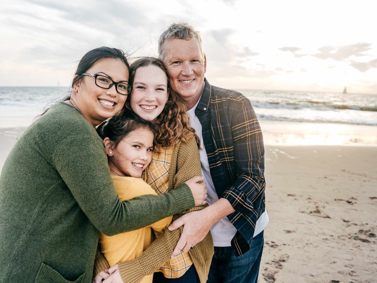 Two parents and two daughters hugging and smiling on a sandy beach at sunset