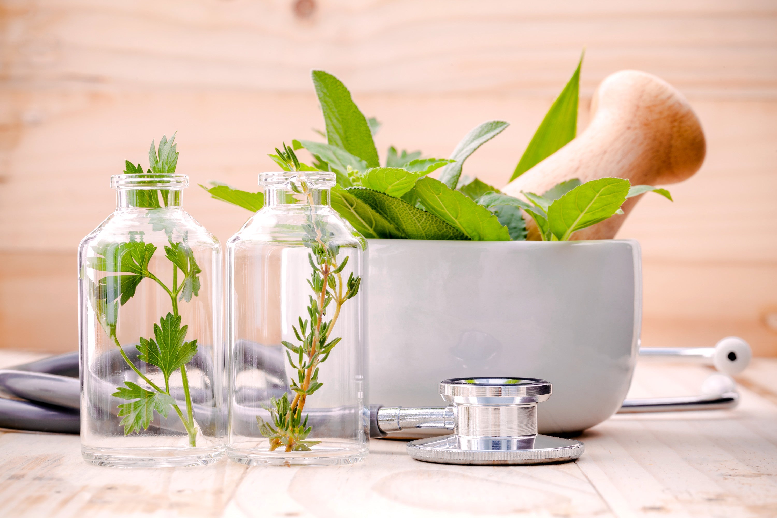 Glass vials with fresh herbal sprigs beside a ceramic mortar and wooden pestle and a stethoscope on a light wood table