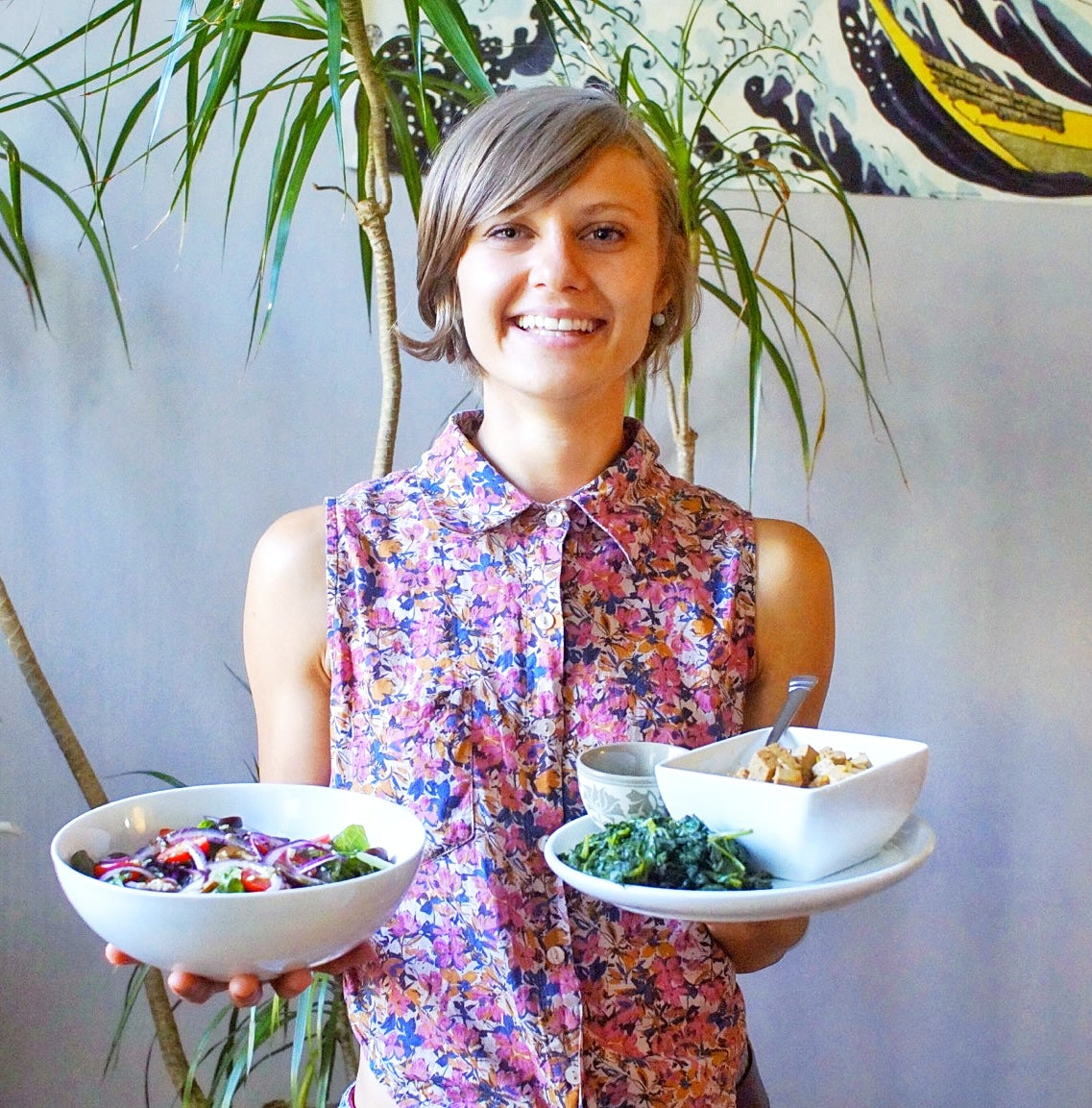 Smiling registered dietitian holding bowls of salad, sauteed greens, and tofu in a bright room with potted plants