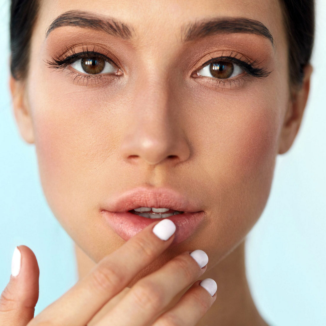 Woman touching her lower lip with her finger in a close-up portrait against a pale blue background, suggesting concern about a cold sore