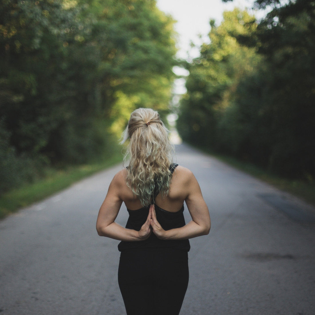 Woman doing reverse prayer yoga pose on a quiet tree-lined road, seen from behind