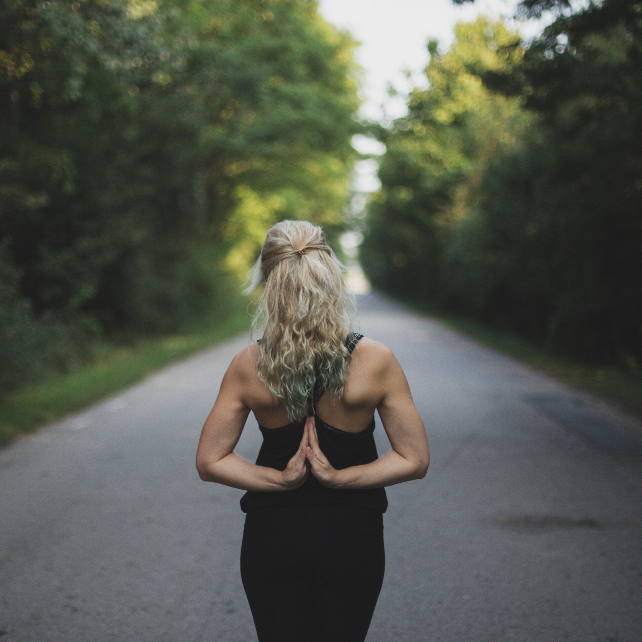 Woman doing reverse prayer yoga pose on a quiet tree-lined road, seen from behind