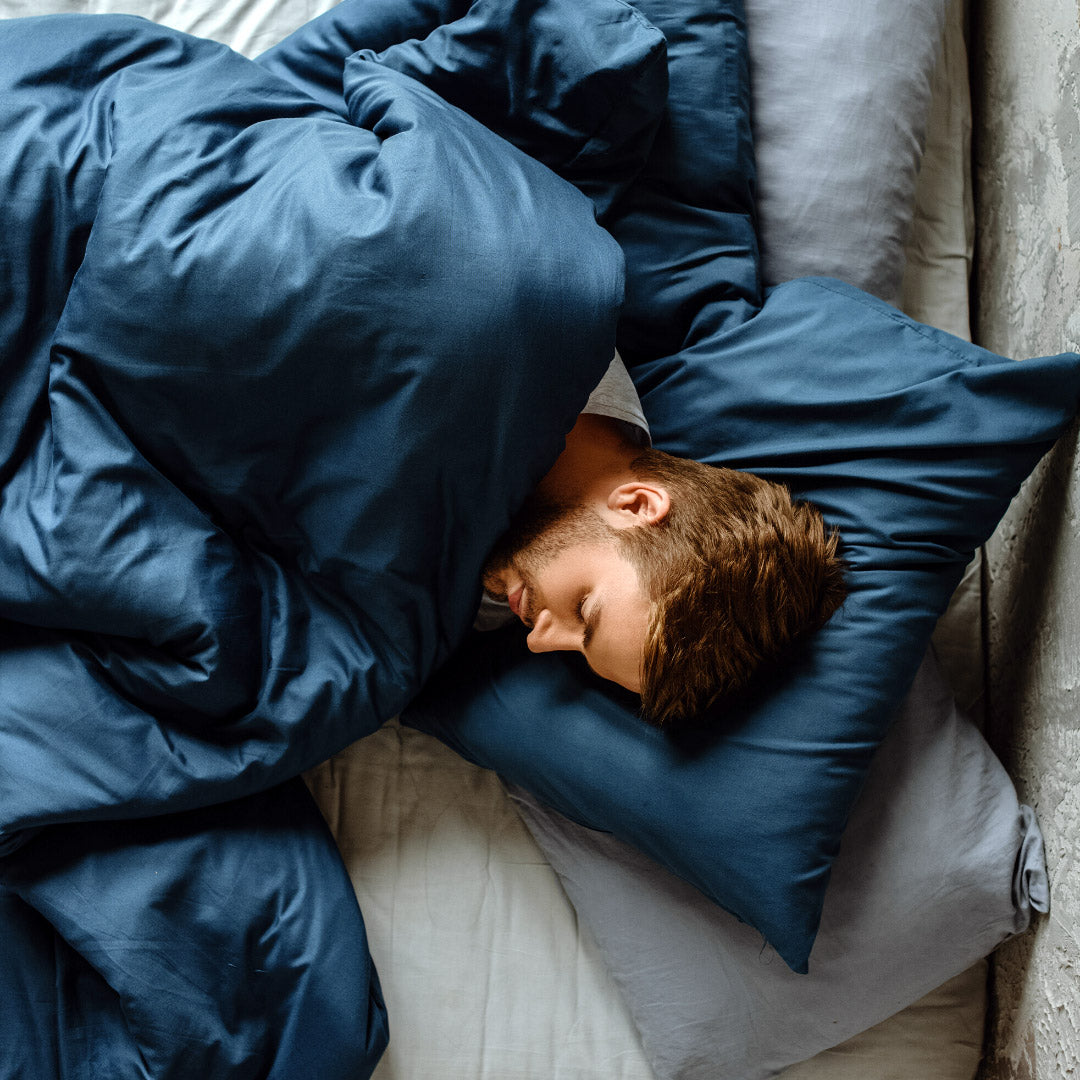 Man sleeping on his side under blue duvet and matching pillows on a bed in a softly lit bedroom