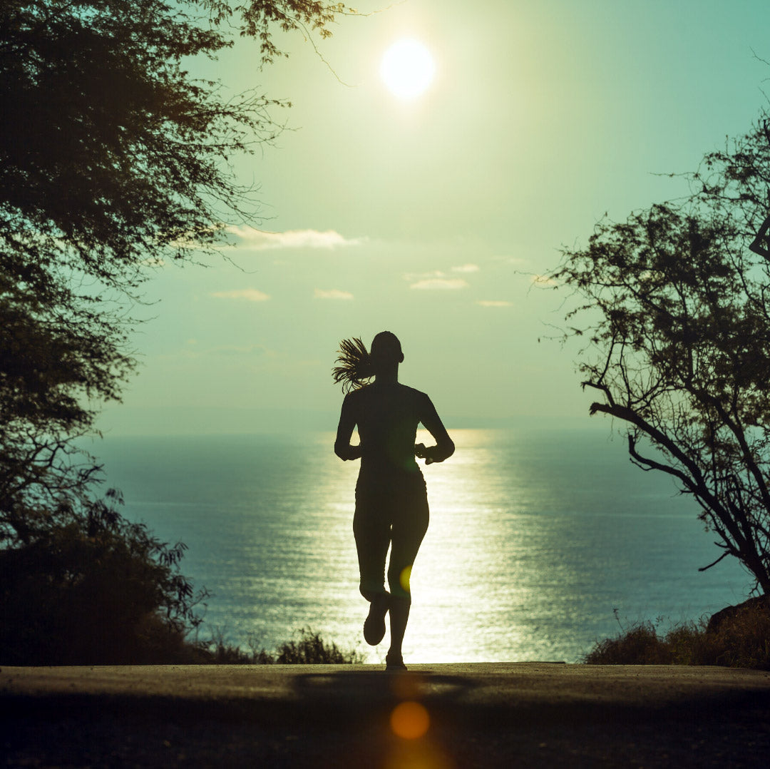 Silhouetted woman jogging toward the ocean on a coastal path backlit by the sun