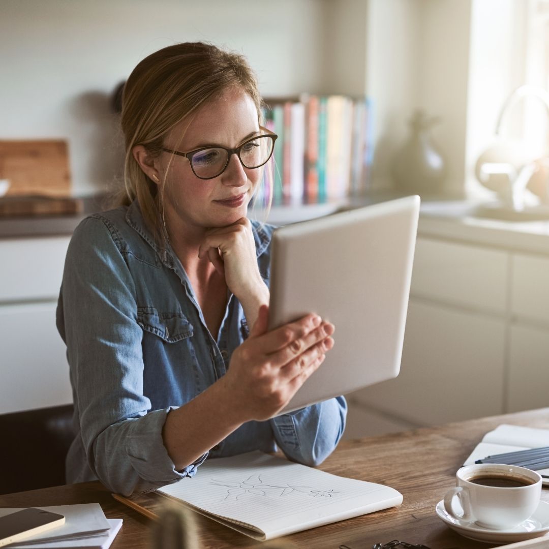 Woman with glasses reading a tablet at a kitchen table with a notebook and a cup of coffee, sunlight and books in the background