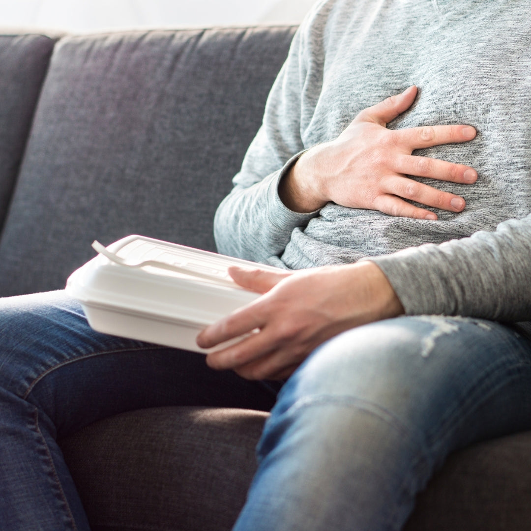 Adult clutching upper abdomen on a couch while holding a closed takeout container, suggesting heartburn or indigestion