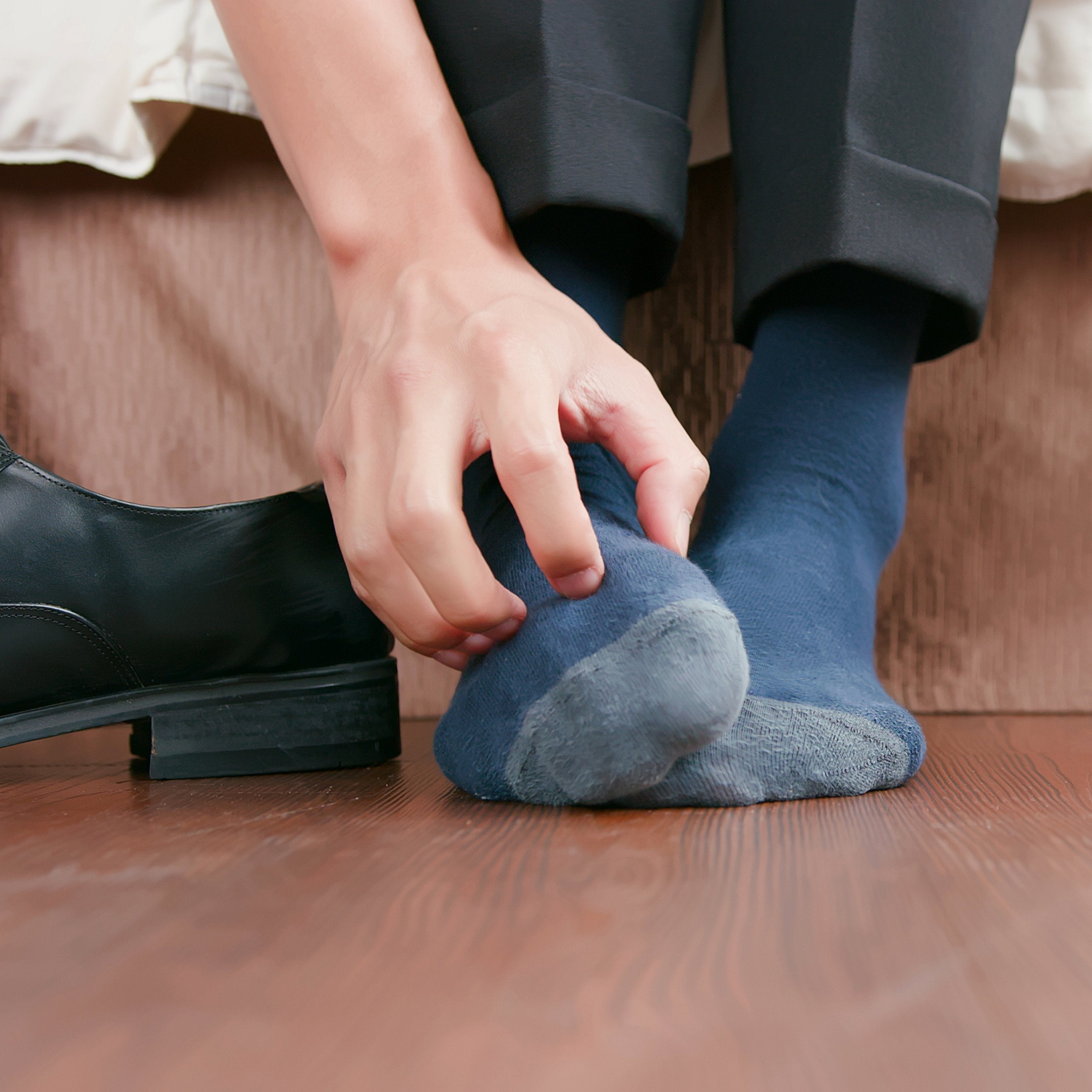 Close-up of a person pulling on a blue sock while sitting on a bed with a black dress shoe nearby