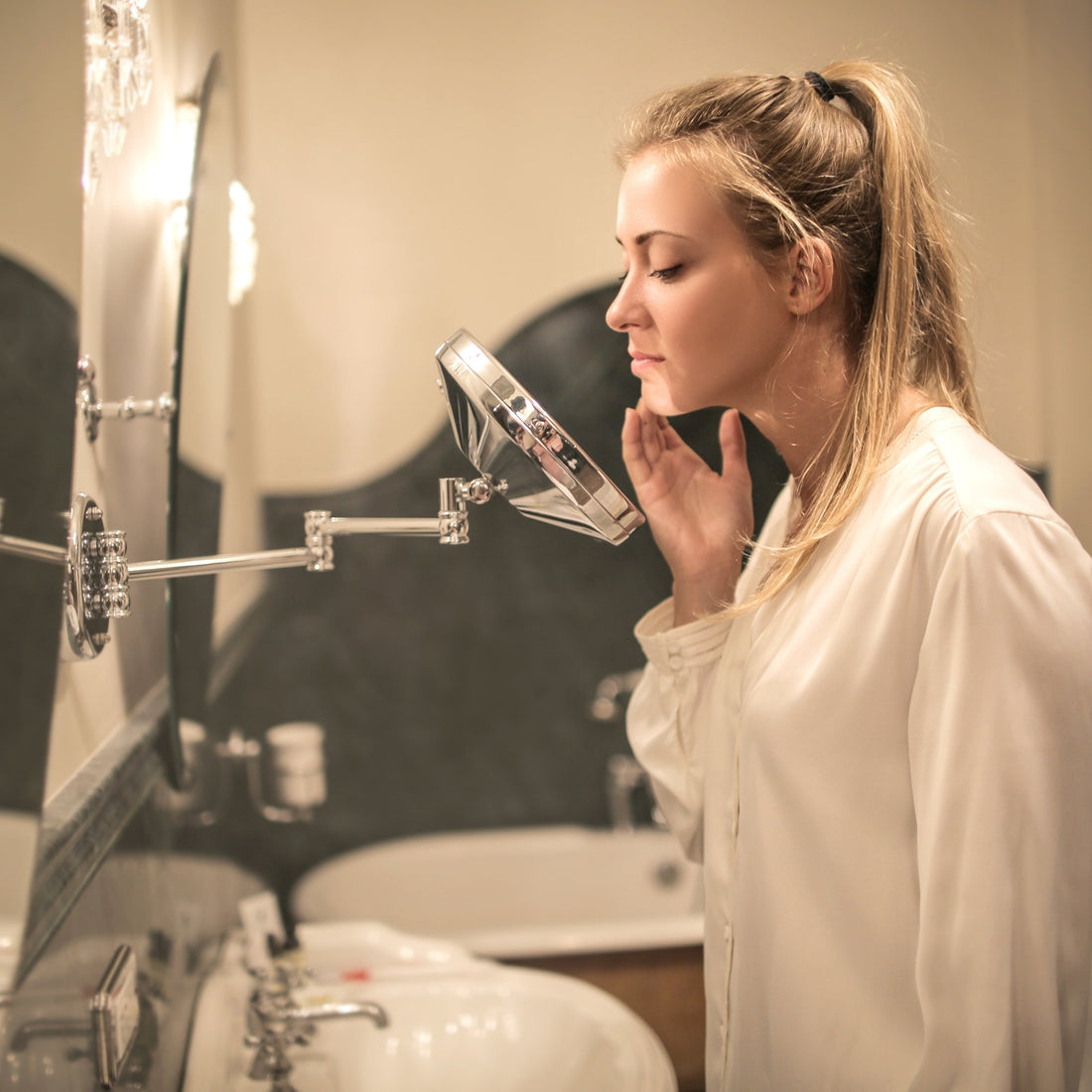 Woman examining her facial skin in a handheld mirror at a bathroom sink, checking for acne