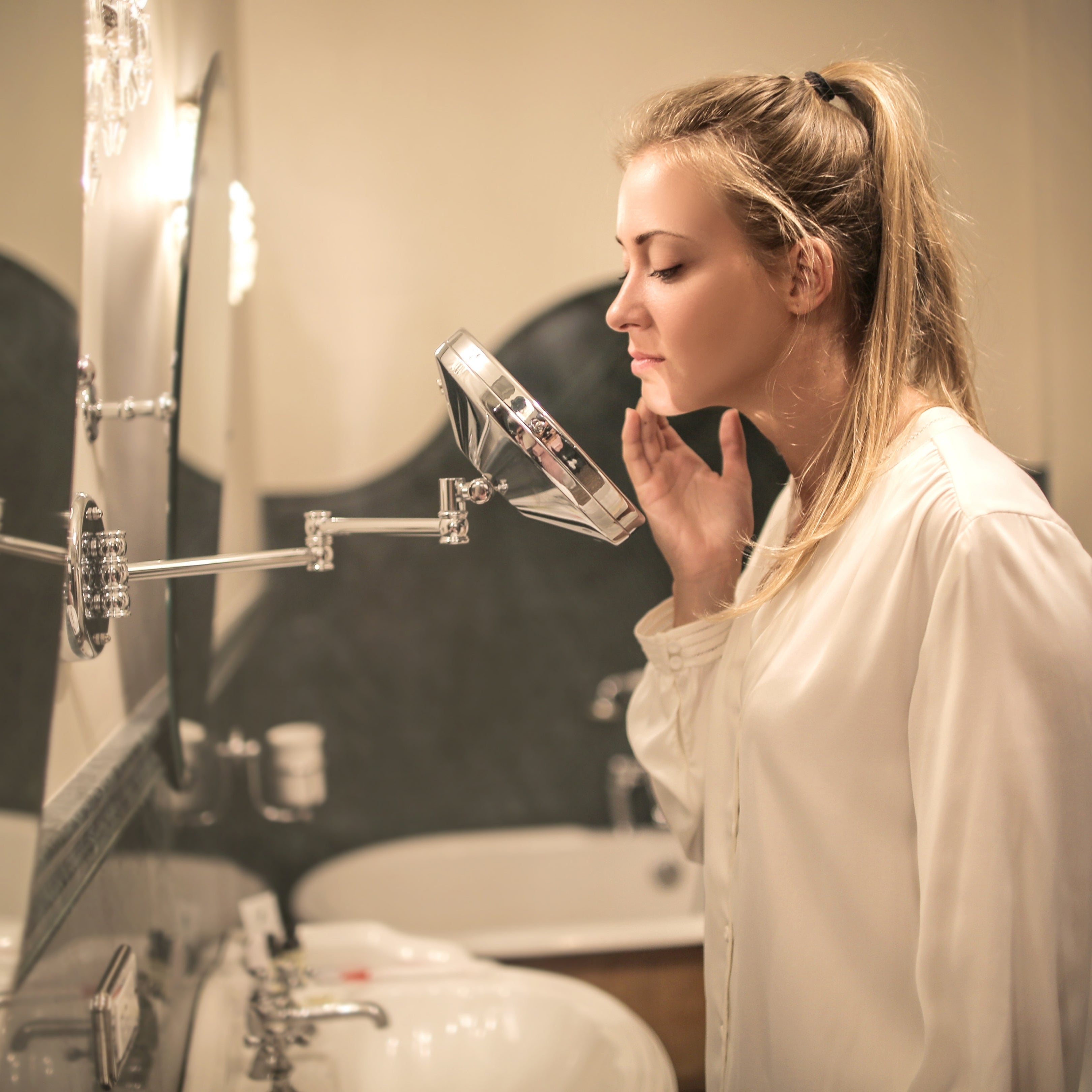 Woman examining her facial skin in a handheld mirror at a bathroom sink, checking for acne