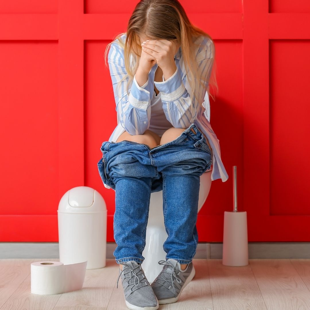 Woman sitting on a toilet with jeans pulled down, clutching her face in distress against a red wall, toilet paper nearby