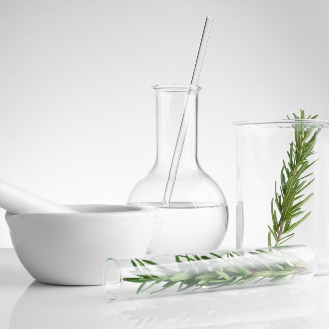 Mortar and pestle with glass beakers and a test tube of rosemary on a white tabletop, natural medicine concept