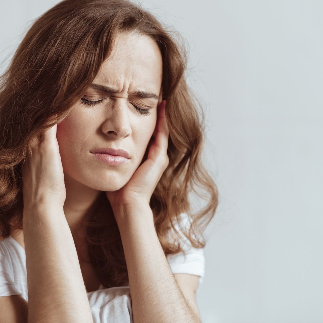 Woman clutching her temples with eyes closed, appearing to have a migraine against a neutral gray background