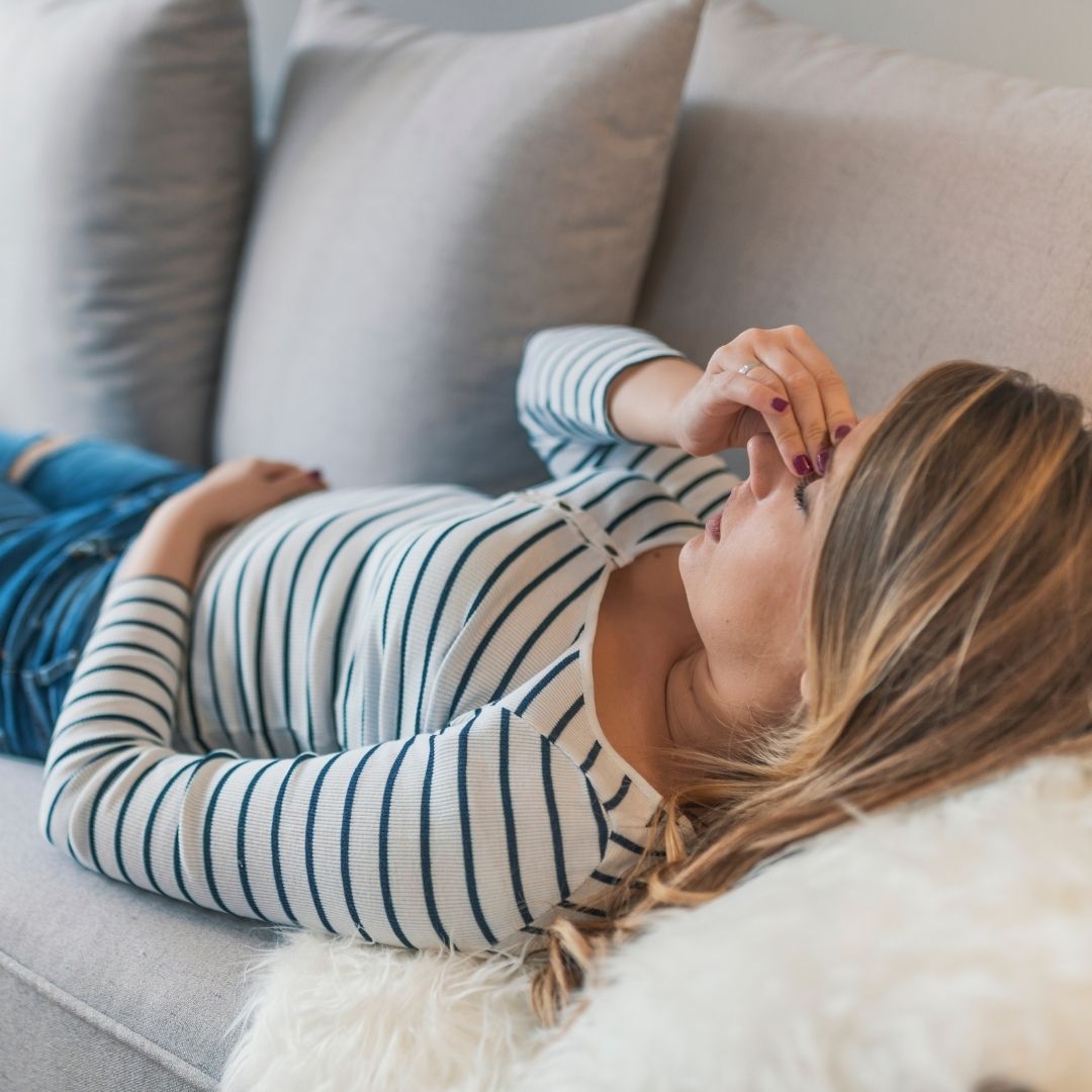 Woman lying on a couch holding the bridge of her nose and rubbing her eyes, appearing to have a headache