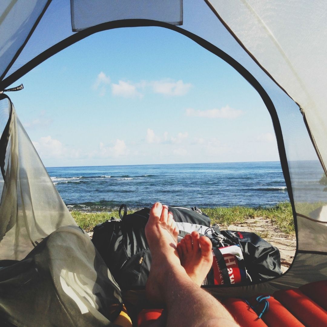 Camper’s bare feet resting inside a tent opening facing a calm ocean and sandy shoreline