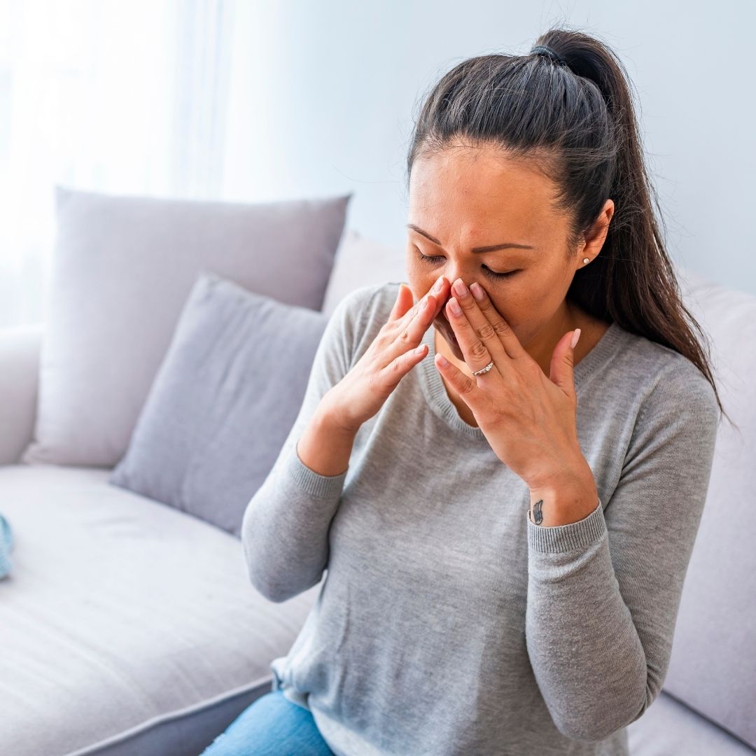 Woman on a couch pressing the bridge of her nose, appearing to have sinus pain and nasal congestion