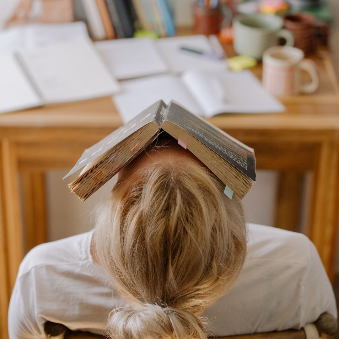 Blond-haired person leaning back with an open book resting on their head at a cluttered study desk