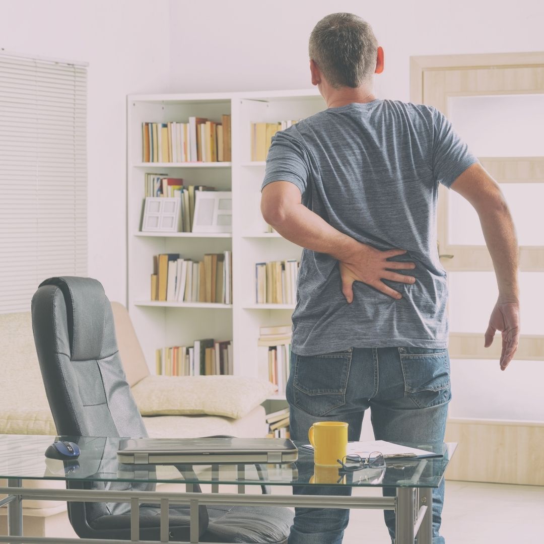Middle-aged man clutching his lower back in pain while standing in a home office beside a glass desk with laptop and yellow mug