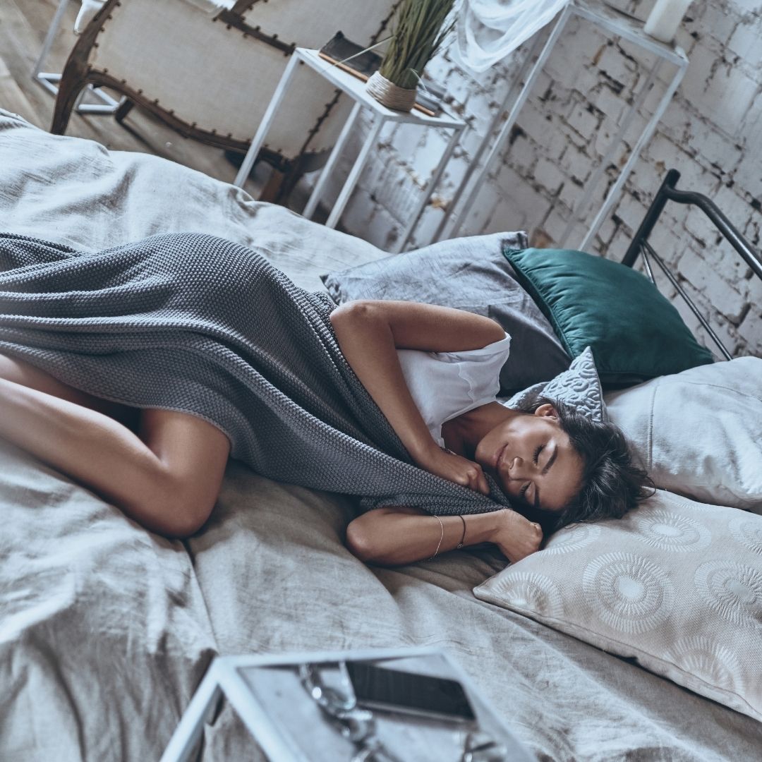 Woman sleeping on bed wrapped in a gray knit blanket in a sunlit loft-style bedroom
