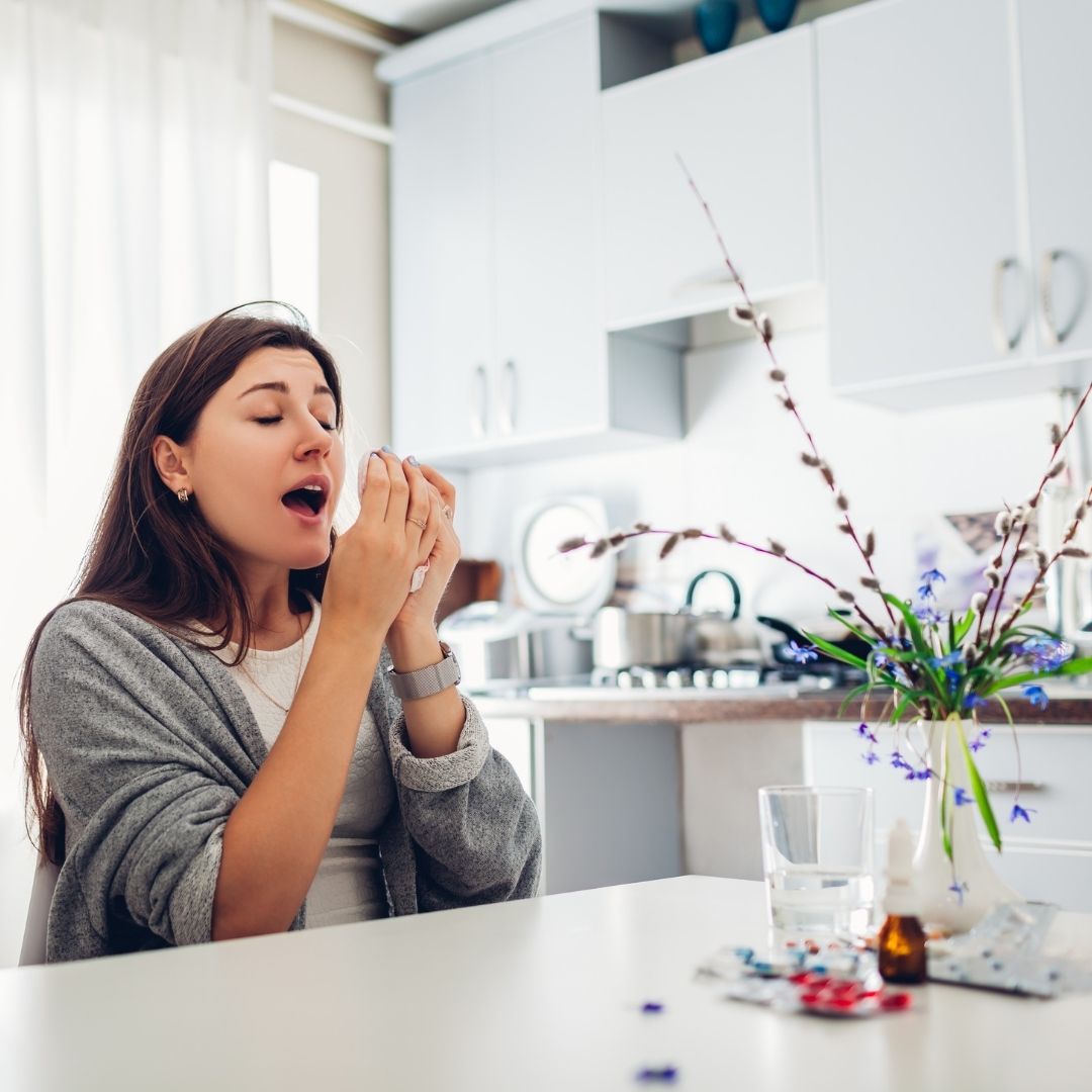 Woman sneezing at a kitchen table next to a vase of flowers and allergy medicine