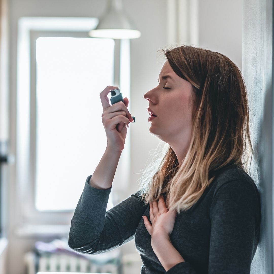 Woman using an asthma inhaler while holding her chest in a bright room